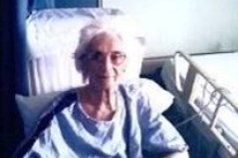 A photo of Alice Brennan sitting beside a hospital bed. She appears in a blue patient gown. 
