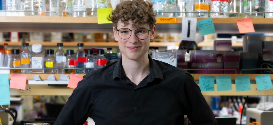 Student in a lab standing in front of shelves with glass bottles. 