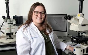 Biochemistry student next to microscopes in a lab coat. 