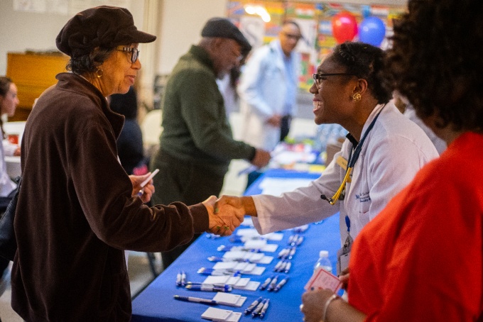 Medical student in a white coat shaking hands with a visitor at a table. 