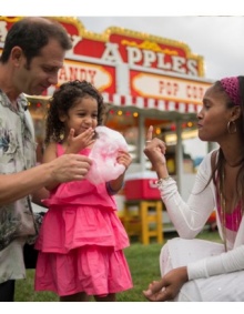 Two adults with a child eating cotton candy in front of a food stand. 