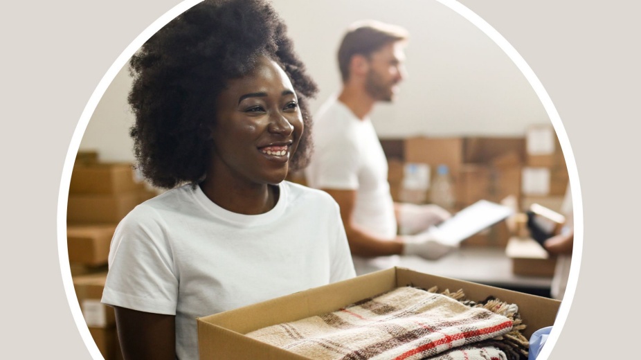 Black woman handing a box full of blankets, a white man is blurred in the background. 