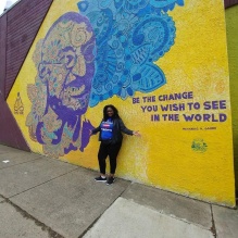 Evette Phillips standing up in a big yellow mural with Ghandi's portrait in purple. Reads "Be the change you wish to see in the world," Mohandas K. Gandhi. 
